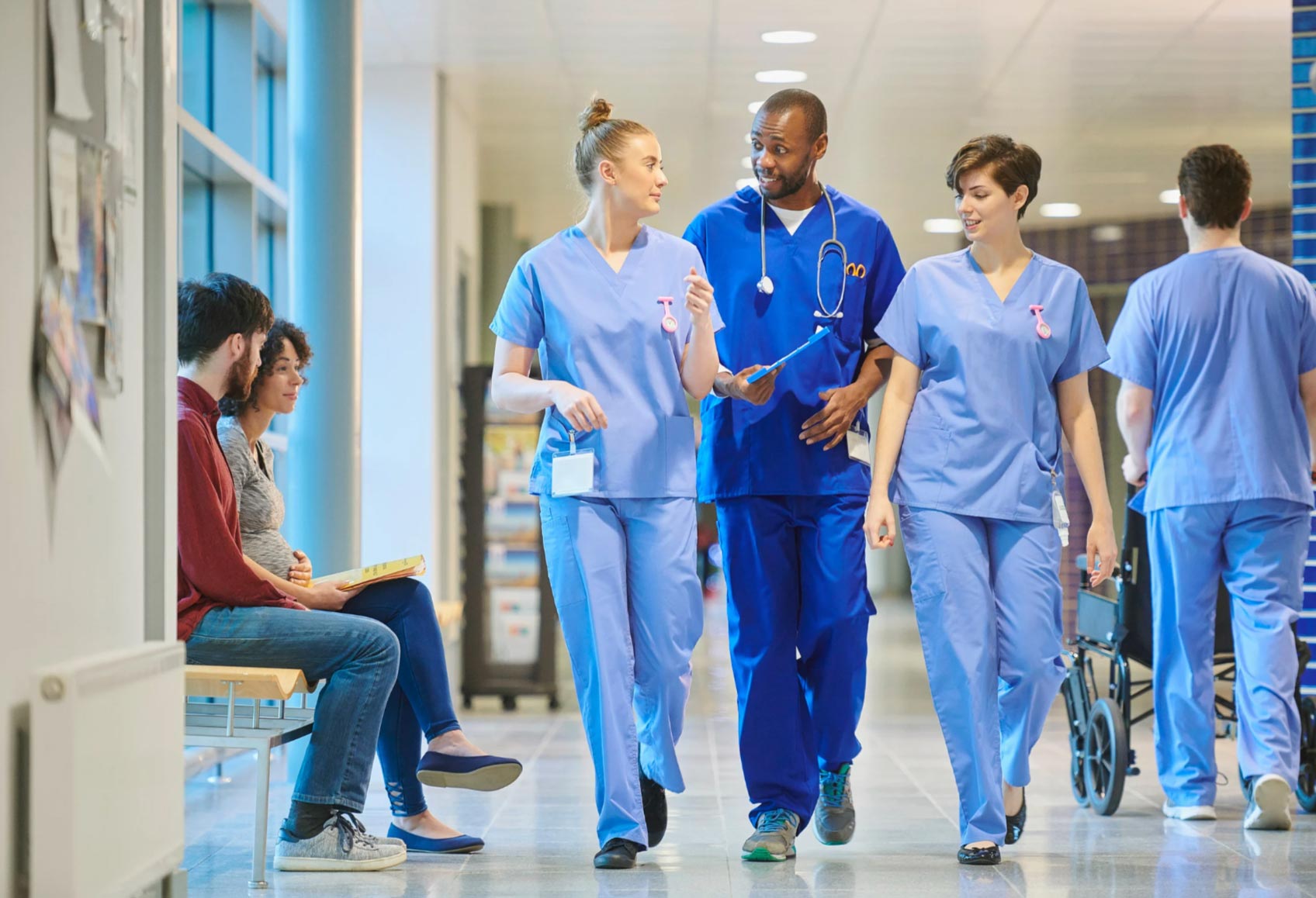 Three medical professionals walk and talk in a hospital hallway while two people sit on a bench nearby.