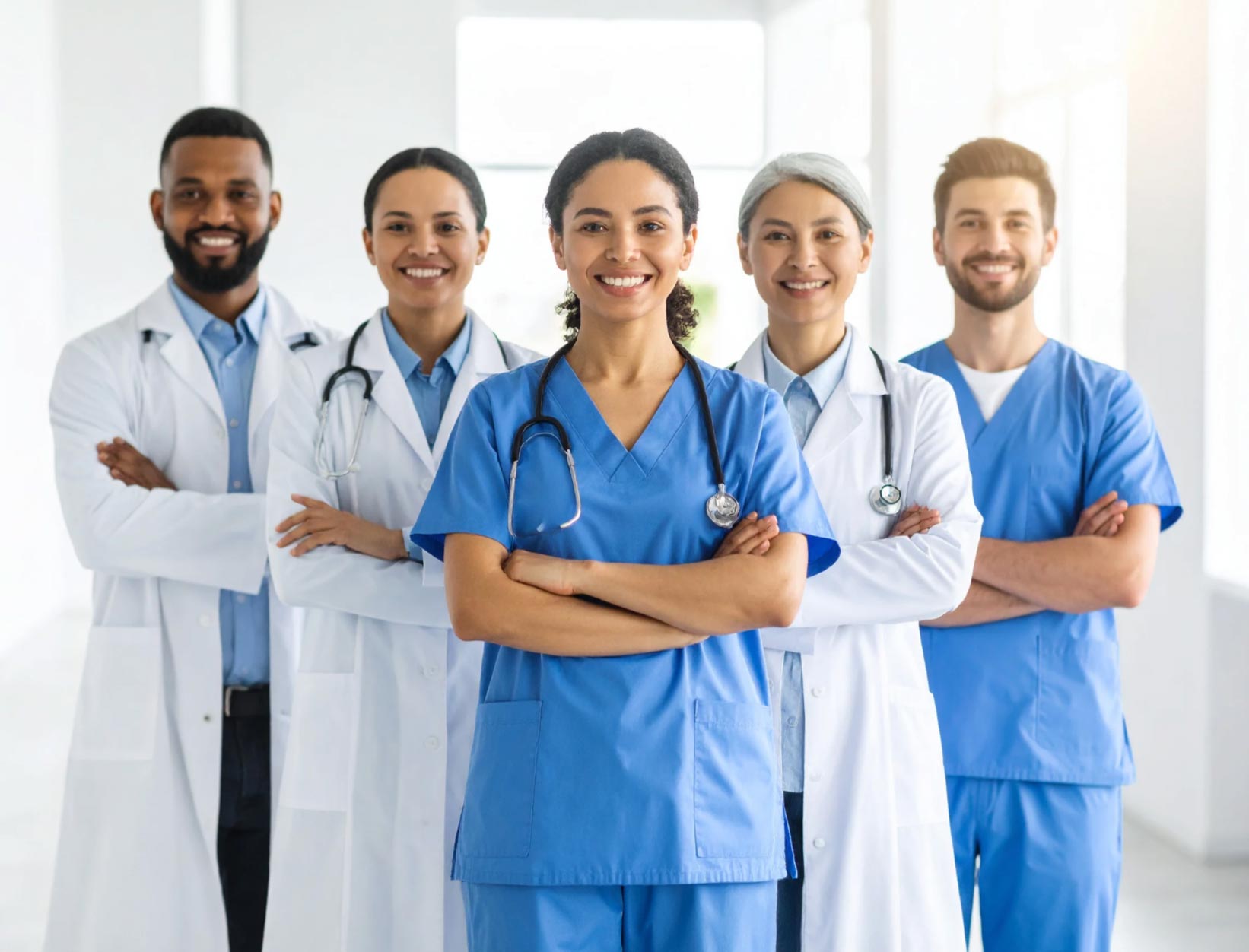 Five smiling healthcare professionals in scrubs and lab coats stand together with arms crossed in a bright clinic.