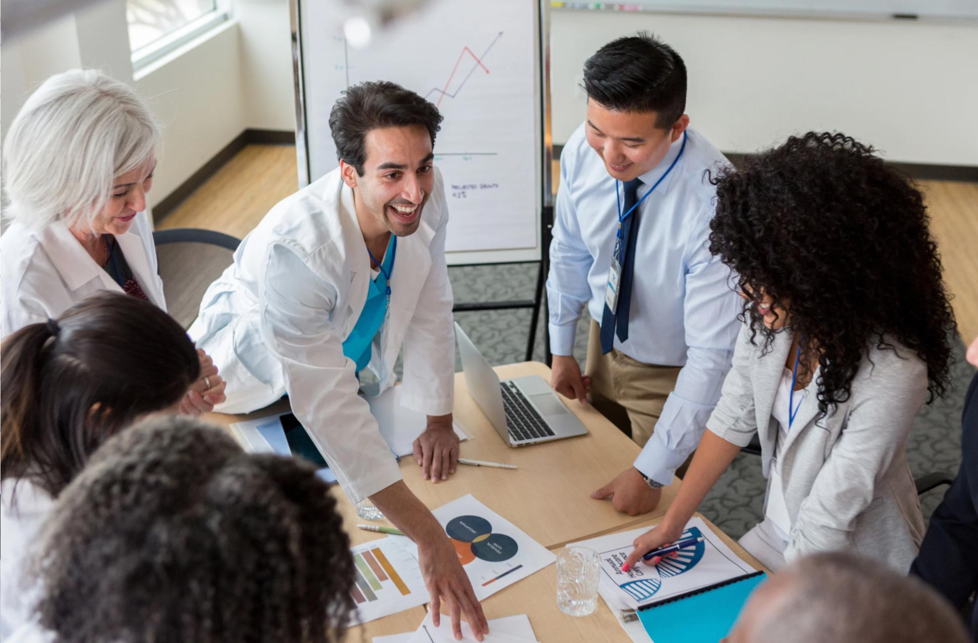 A diverse group of professionals in lab coats and business attire discuss charts around a table.