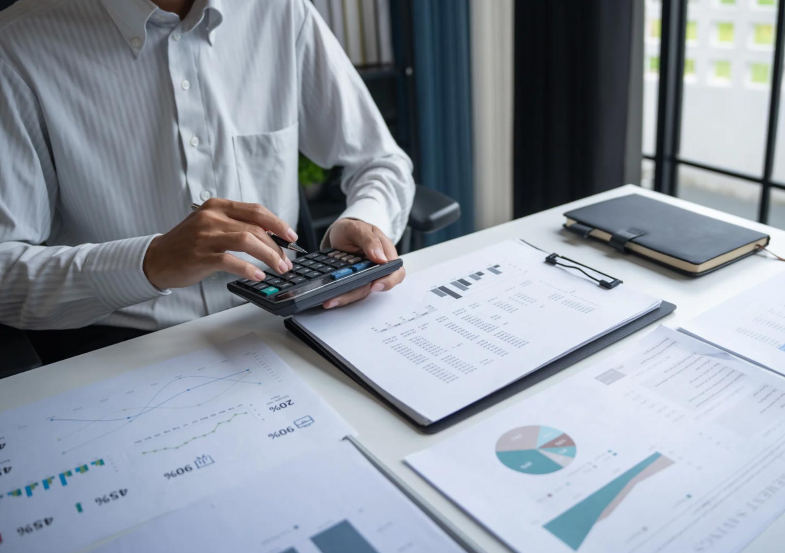 Person using a calculator at a desk with financial charts, graphs, reports, a clipboard, and notebooks.