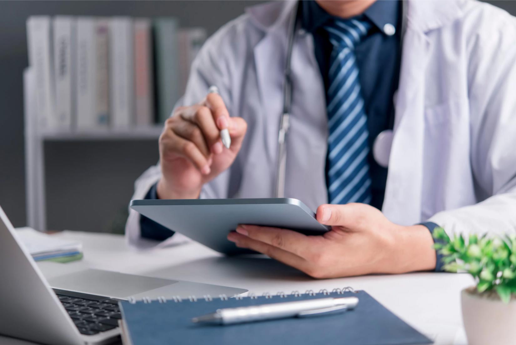 A doctor in a white coat uses a tablet at a desk with a laptop, notebook, and potted plant.