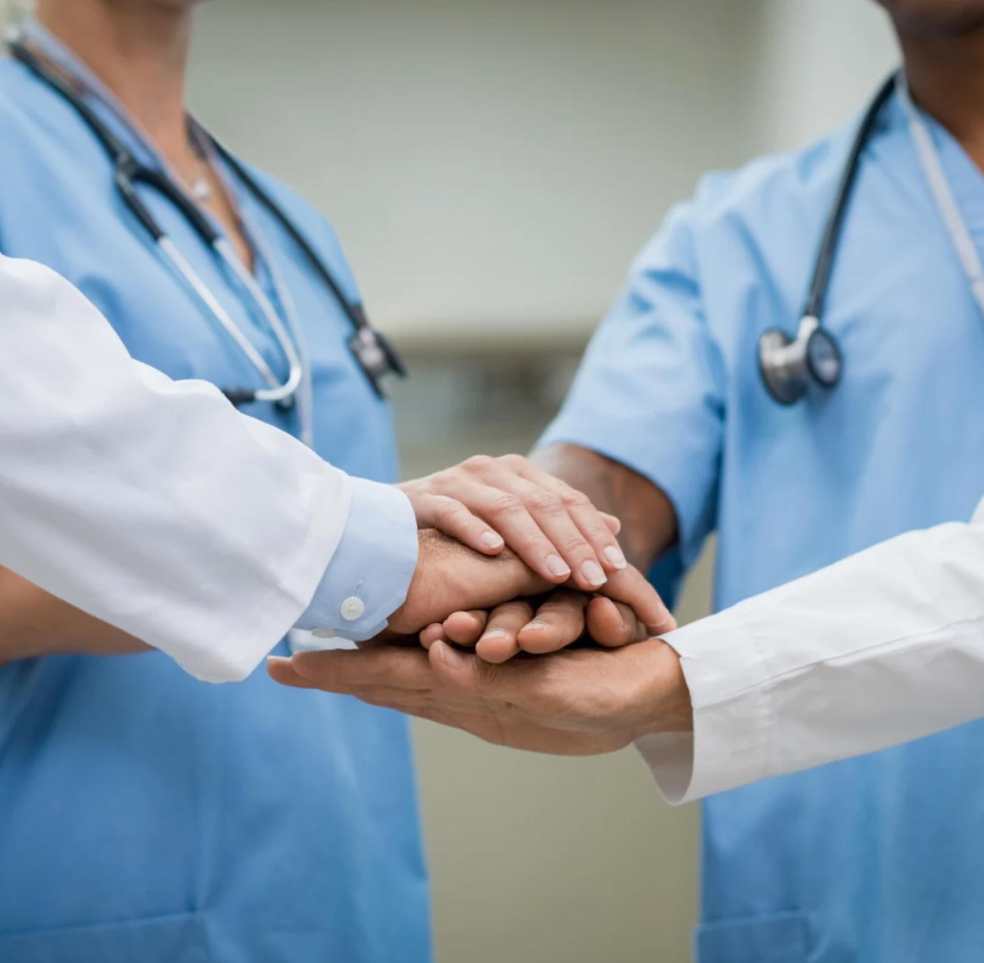 Four healthcare professionals stacking hands in unity, wearing blue scrubs and stethoscopes.