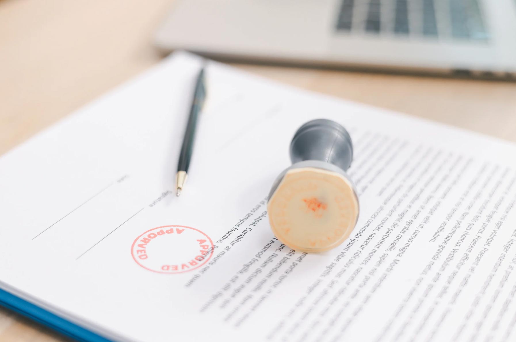 A pen and an "APPROVED" stamp rest on a document beside a laptop.