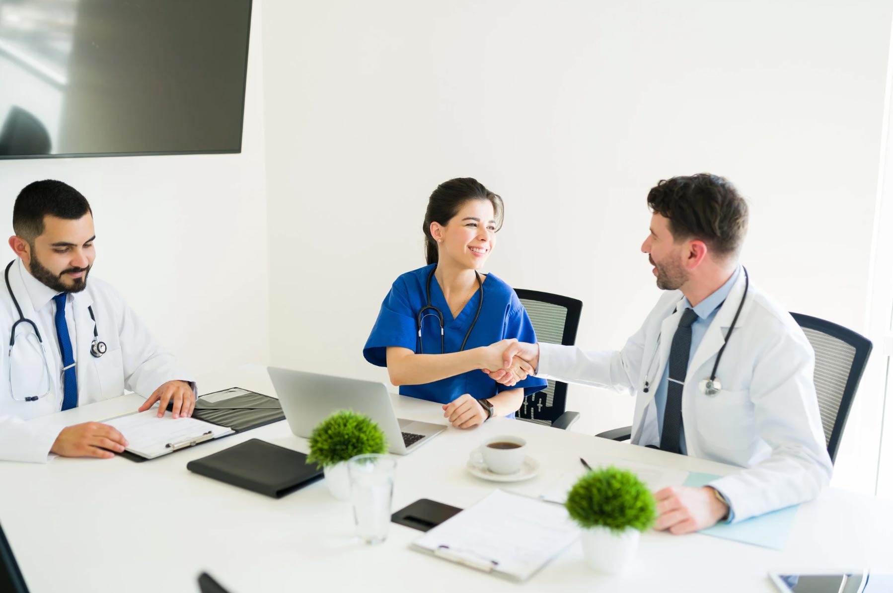 hospital-img Three doctors sit at a table, two shaking hands and smiling, with laptops and documents in front of them.